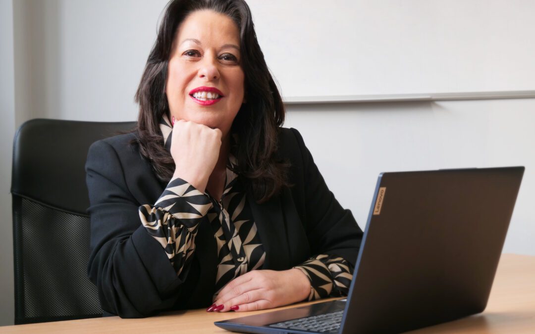 A dark haired woman sits at a desk with a laptop open in front of her.